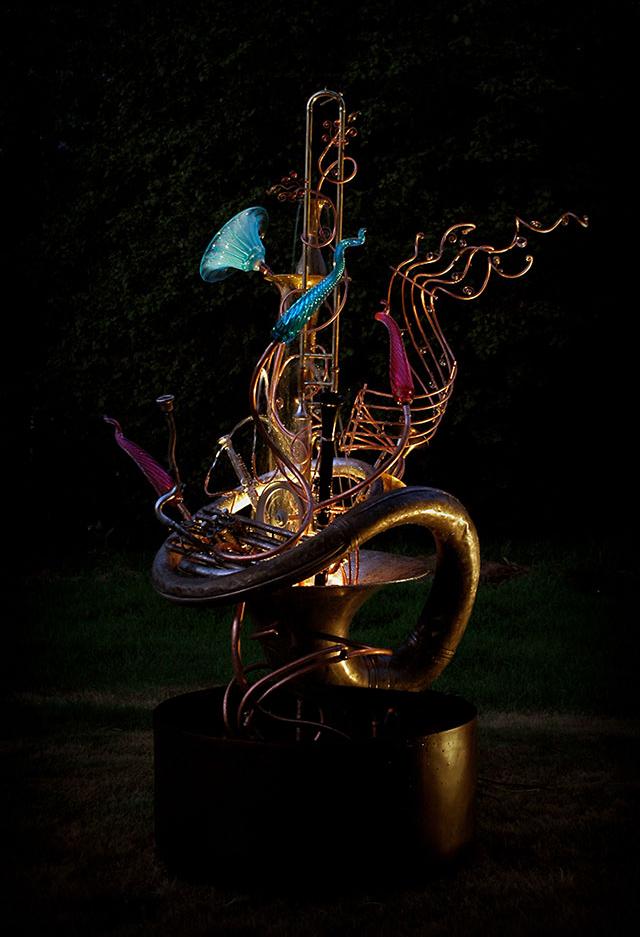 Musical instrument fountain sculpture created from copper tubing, a water wheel, blown glass, a musical scroll, repurposed brass and copper, and upcycled musical instruments including a sousaphone, a sax and a trumpet.
