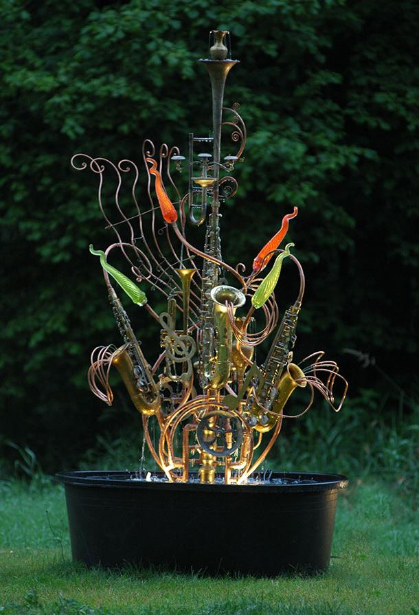 Musical instrument fountain sculpture made from copper tubing, a copper musical scroll, a water wheel, blown glass, recycled brass objects, and upcycled musical instruments including a sousaphone, a sax, and a trumpet.